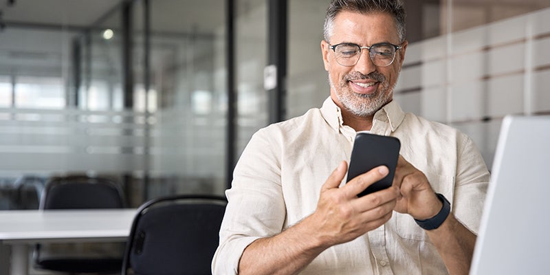 Smiling man in glasses and a button-up shirt looking at his phone in an office.